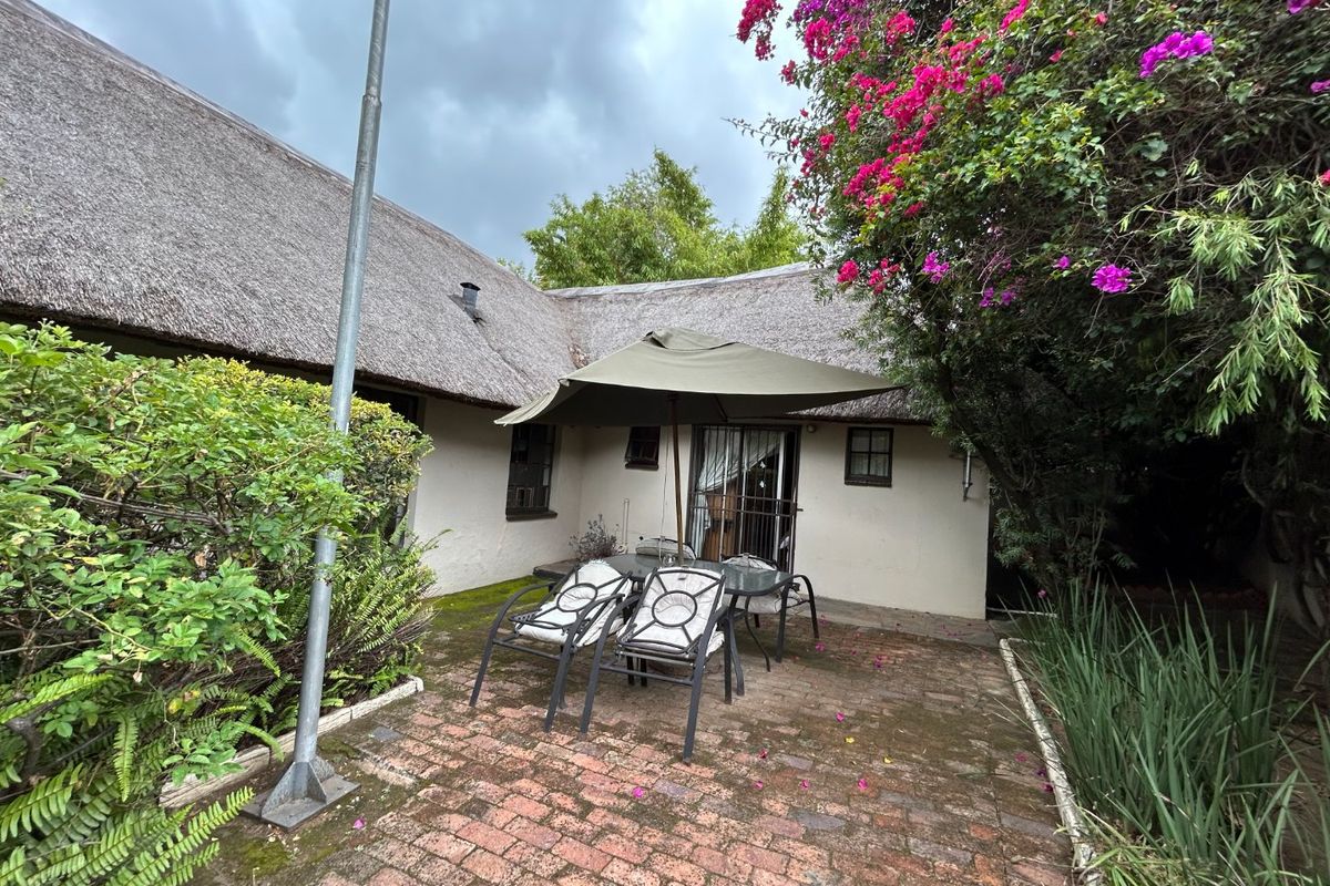 A rear patio area featuring a traditional thatch roof, brick paving, and established garden surroundings with security gate access to the building interior.