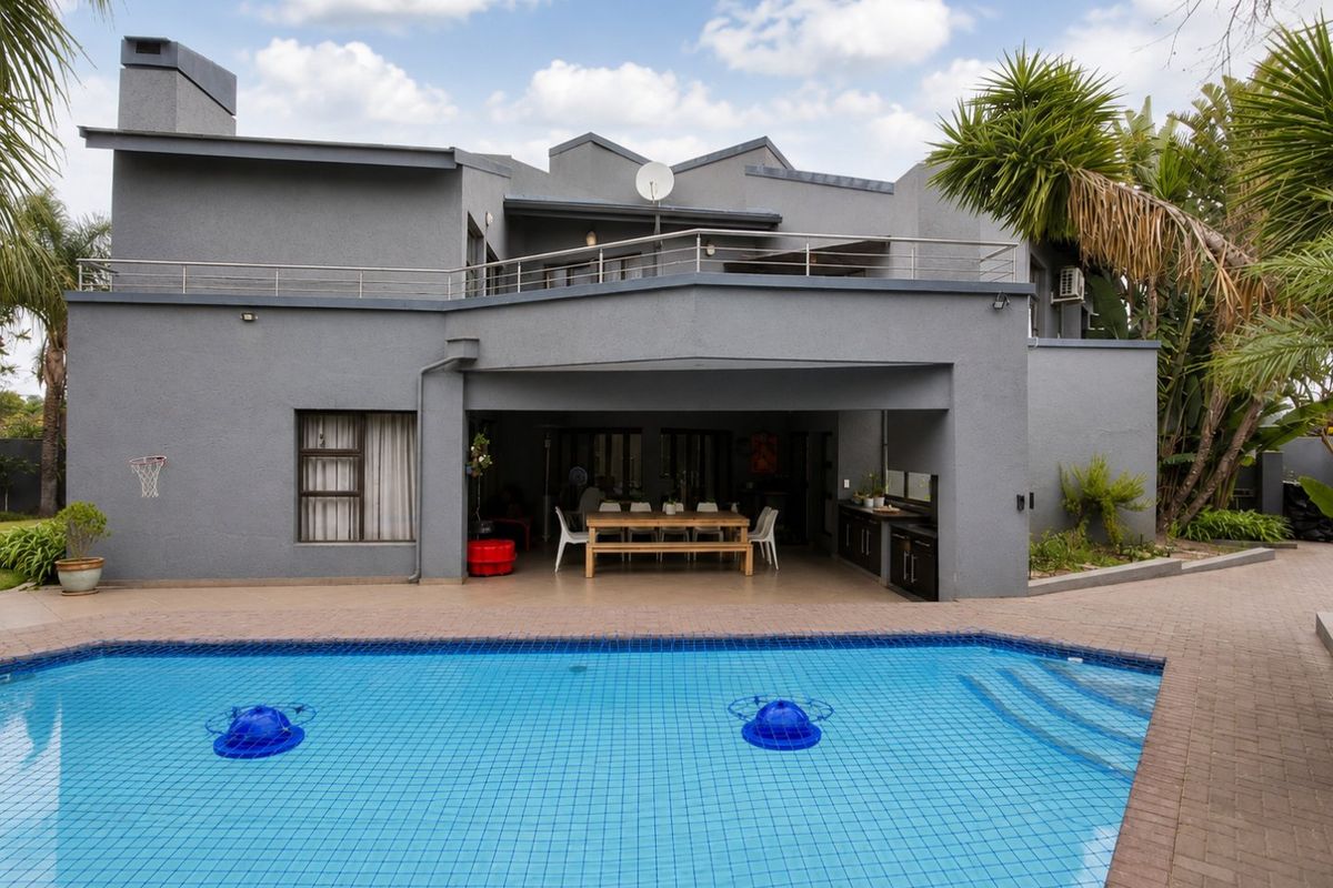 Rear view of a modern grey house featuring a swimming pool, paved deck, covered patio with built-in cabinetry, and an upper-level balcony.