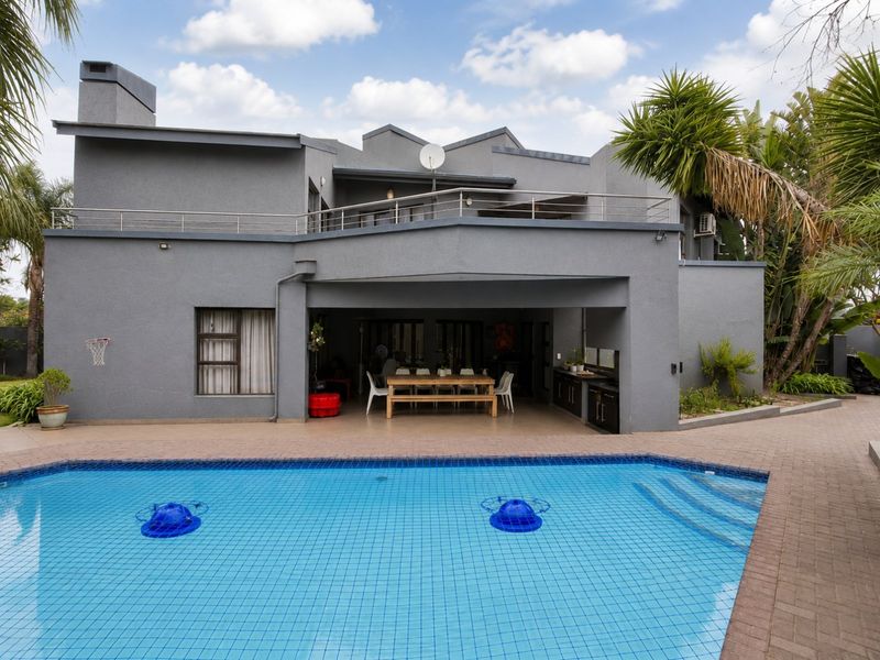 Rear view of a modern grey house featuring a swimming pool, paved deck, covered patio with built-in cabinetry, and an upper-level balcony.