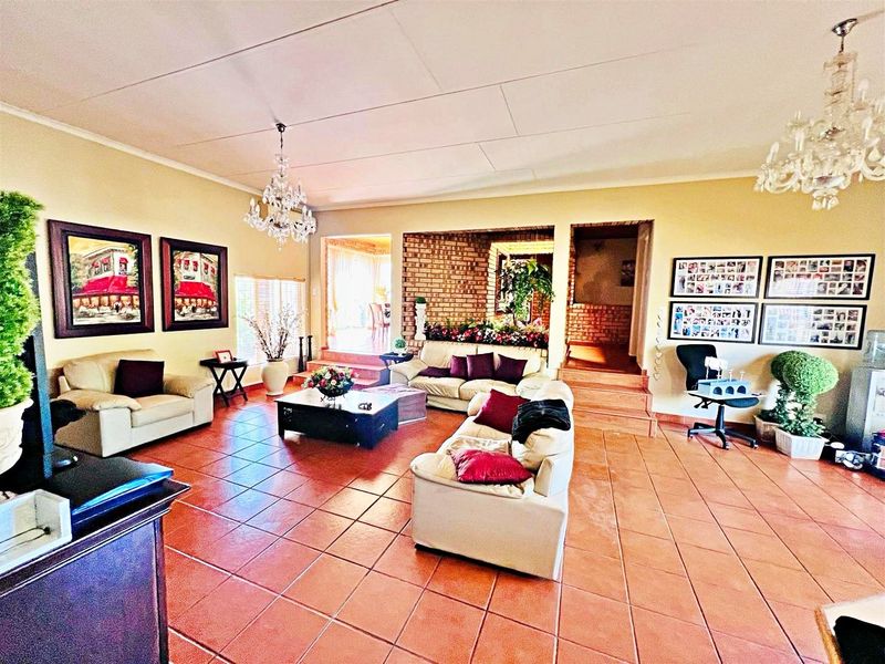 Spacious living room featuring terracotta tiled flooring, an exposed brick feature wall, and elegant crystal chandeliers, with a raised platform area leading to adjacent spaces.