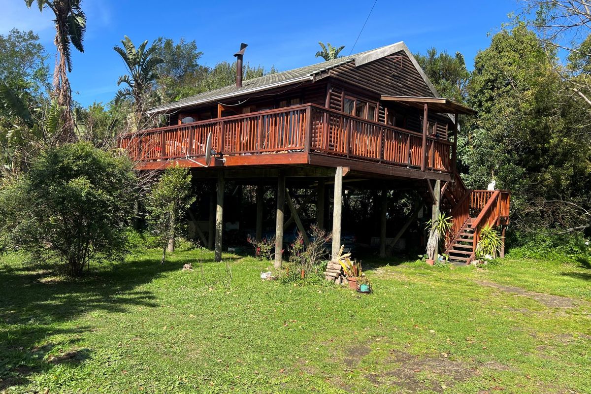 A rustic timber house elevated on stilts, featuring a large wrap-around wooden deck and an external staircase set within a natural garden environment.