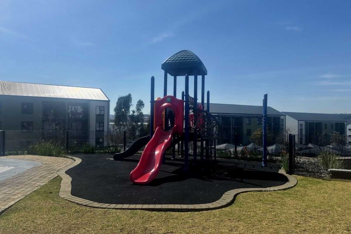 Communal outdoor area featuring a playground with slide and climbing structure, set within a landscaped garden with paved pathways and secure fencing.