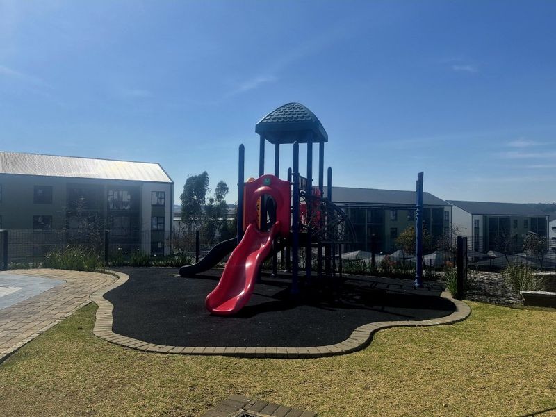 Communal outdoor area featuring a playground with slide and climbing structure, set within a landscaped garden with paved pathways and secure fencing.
