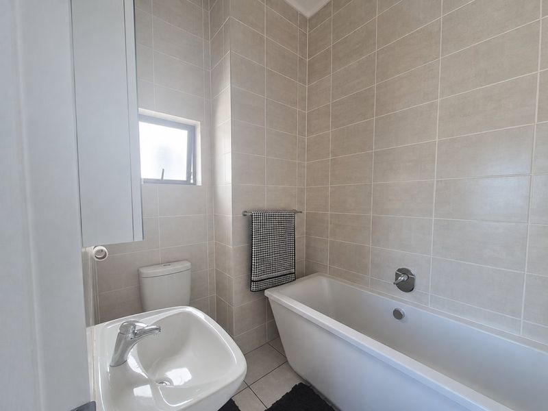 A clean, modern bathroom featuring neutral wall tiles, a white bathtub with wall-mounted fixtures, a pedestal basin, and a toilet.