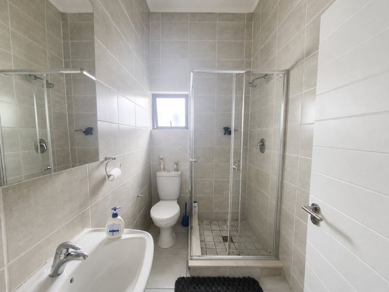 A clean, modern bathroom featuring a glass-enclosed shower, a white toilet, a wall-mounted basin, and neutral floor-to-ceiling wall tiles.