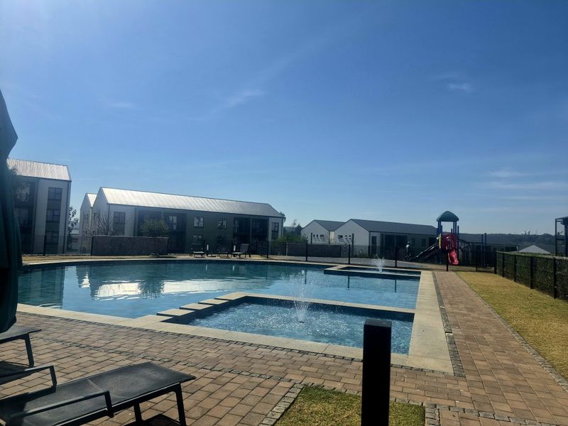 A well-maintained communal swimming pool area featuring a paved deck, integrated water feature, and adjacent playground facilities within the apartment complex.