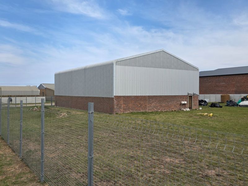Rear view of an industrial warehouse building featuring corrugated metal cladding, a brick base, and surrounding wire mesh perimeter fencing.