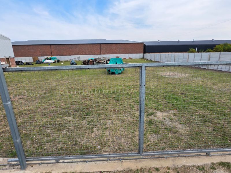 A fenced industrial yard area featuring a precast concrete perimeter wall and adjacent metal-clad industrial buildings.