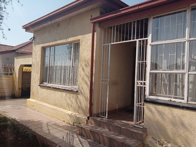 Front exterior view of the house featuring plastered walls, security gate, and tiled entrance steps, showing signs of wear and maintenance requirements.