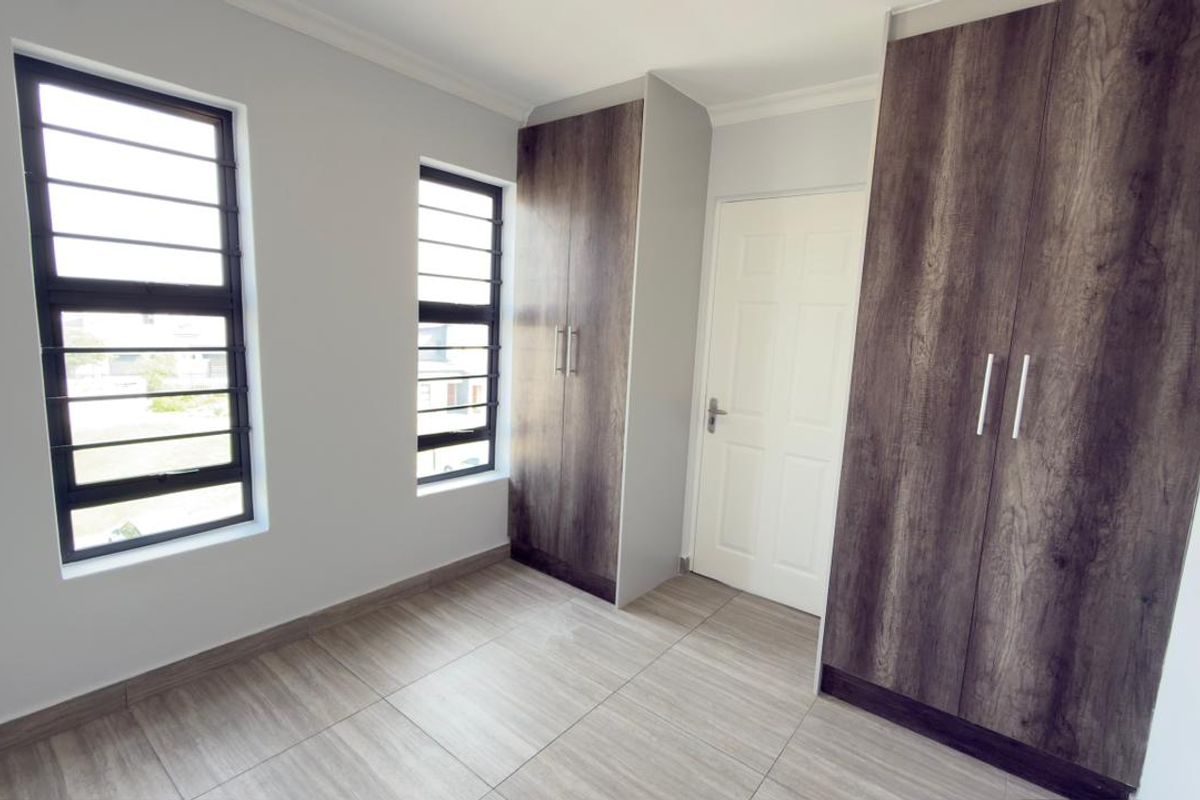 A bright, empty bedroom featuring modern wood-finish built-in cupboards, tiled flooring, and windows fitted with security bars.