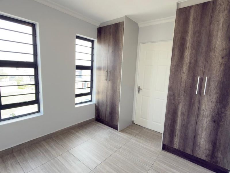 A bright, empty bedroom featuring modern wood-finish built-in cupboards, tiled flooring, and windows fitted with security bars.