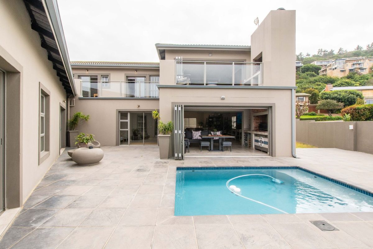 Rear exterior view featuring a swimming pool, tiled patio area, built-in braai, and upper-level balcony with glass balustrades.