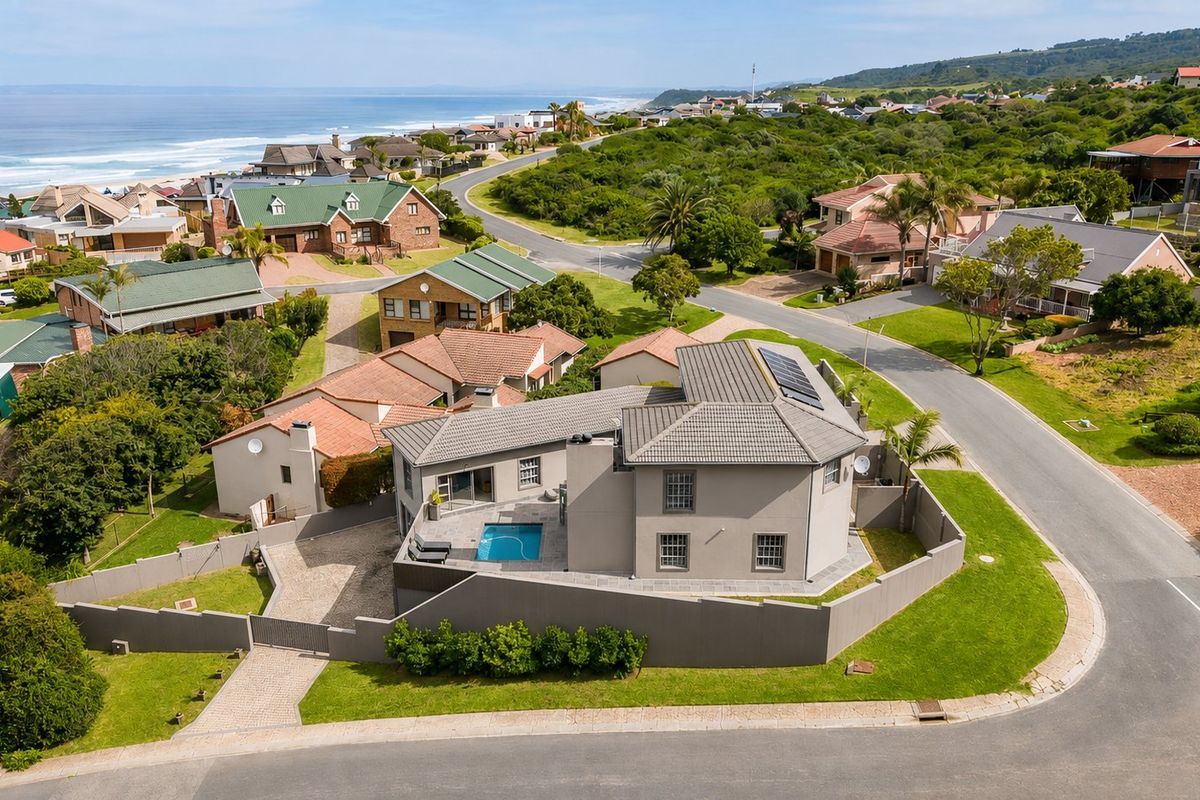 Aerial view of a coastal residential property featuring a swimming pool, solar panels, and a paved driveway within a landscaped estate setting.
