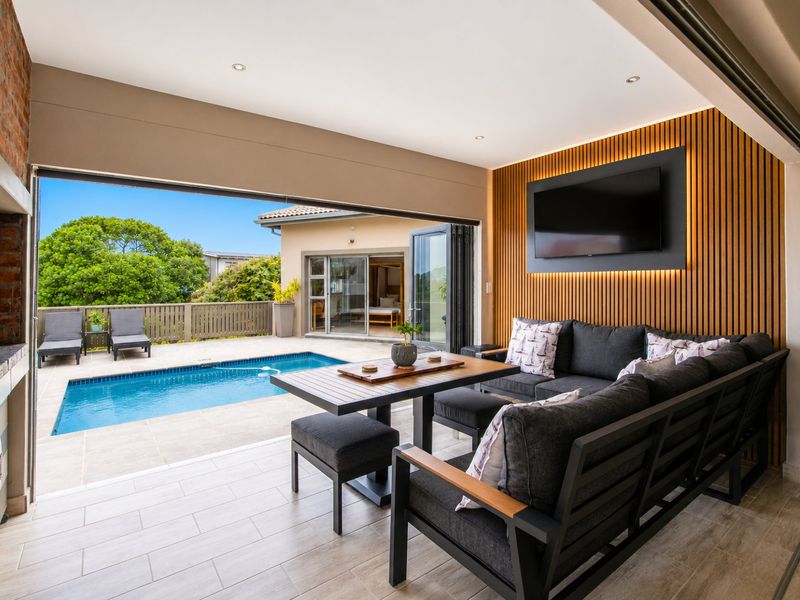 Covered patio featuring a built-in braai, timber slat wall, and seamless flow to the pool area through large stacking doors.