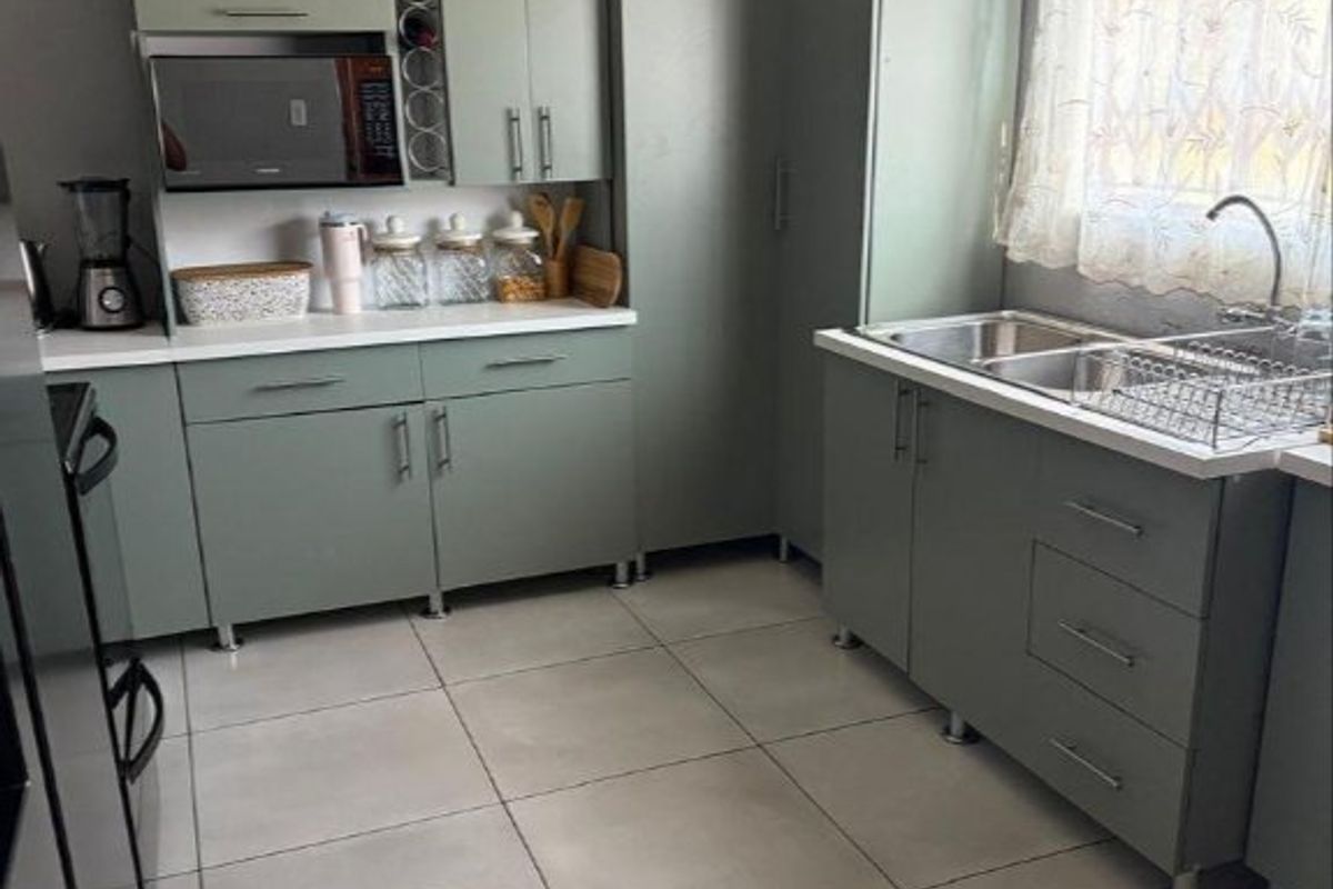 A functional kitchen featuring sage green cabinetry, laminate countertops, a double bowl stainless steel sink, and neutral tiled flooring.