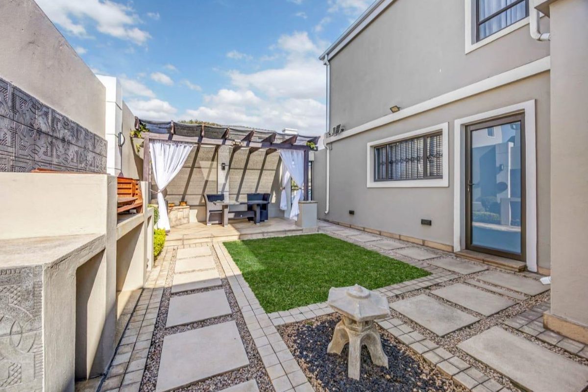 A well-maintained rear courtyard featuring a paved patio, built-in braai area, pergola, and a neat artificial lawn section.