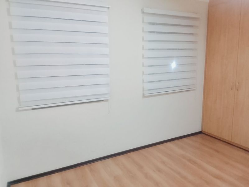 A clean, empty bedroom featuring light wood-look laminate flooring, built-in wooden cupboards, and modern white roller blinds on the windows.