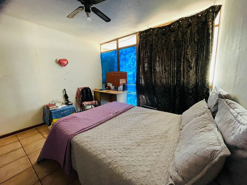A bedroom featuring tiled flooring and a ceiling fan, with large windows providing natural light. The space requires cosmetic updates to the wall finishes.