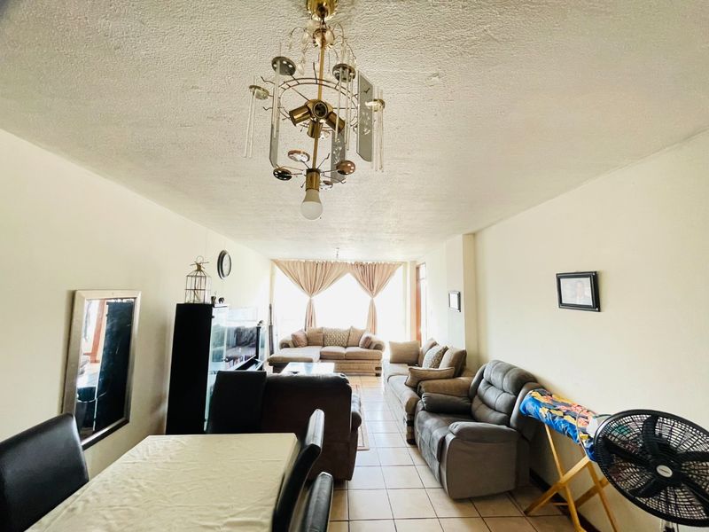 Spacious open-plan living and dining area featuring ceramic tile flooring, a central decorative chandelier, and access to a balcony through sliding glass doors.