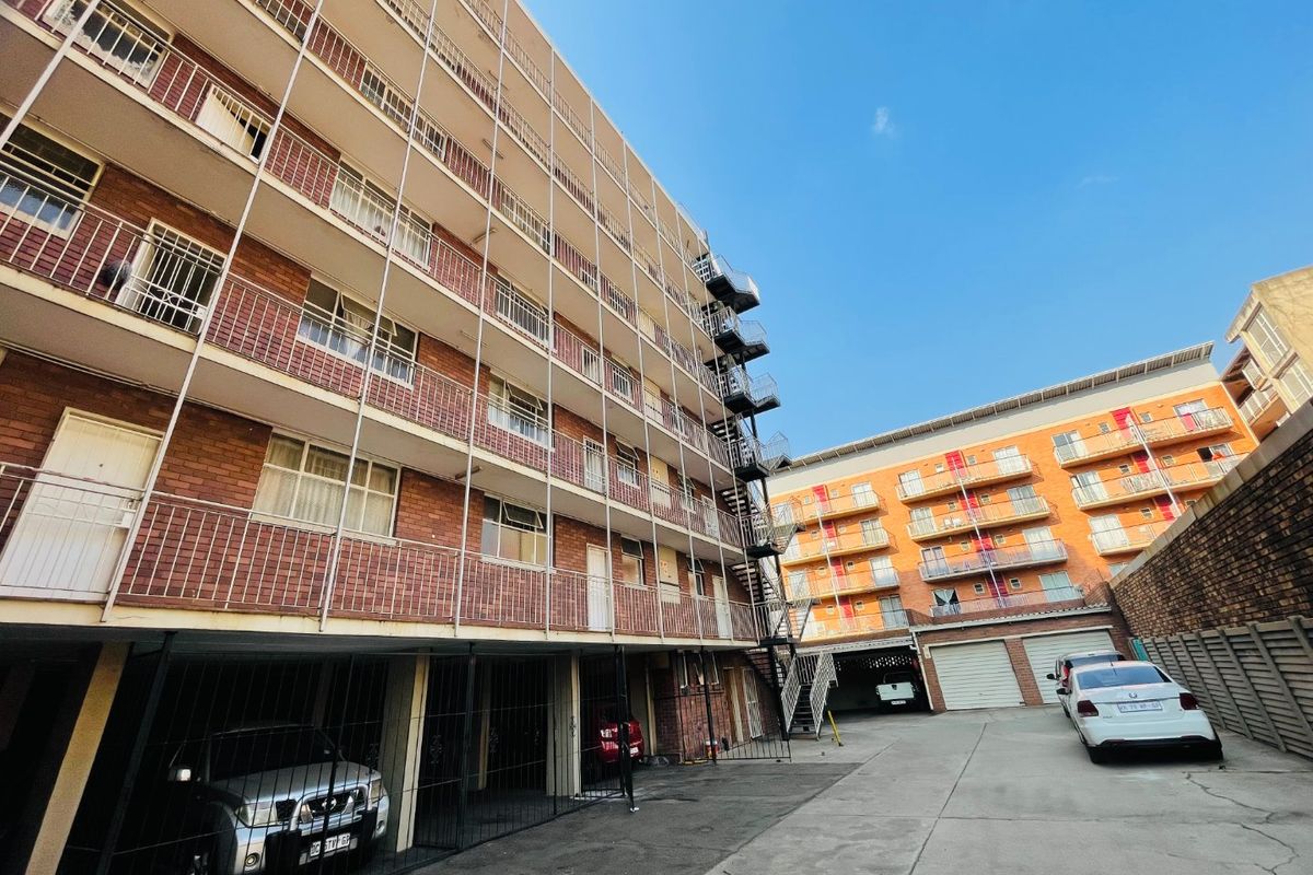Exterior view of a multi-story apartment complex featuring brick facades, metal balconies, a fire escape staircase, and a paved ground-level parking area.