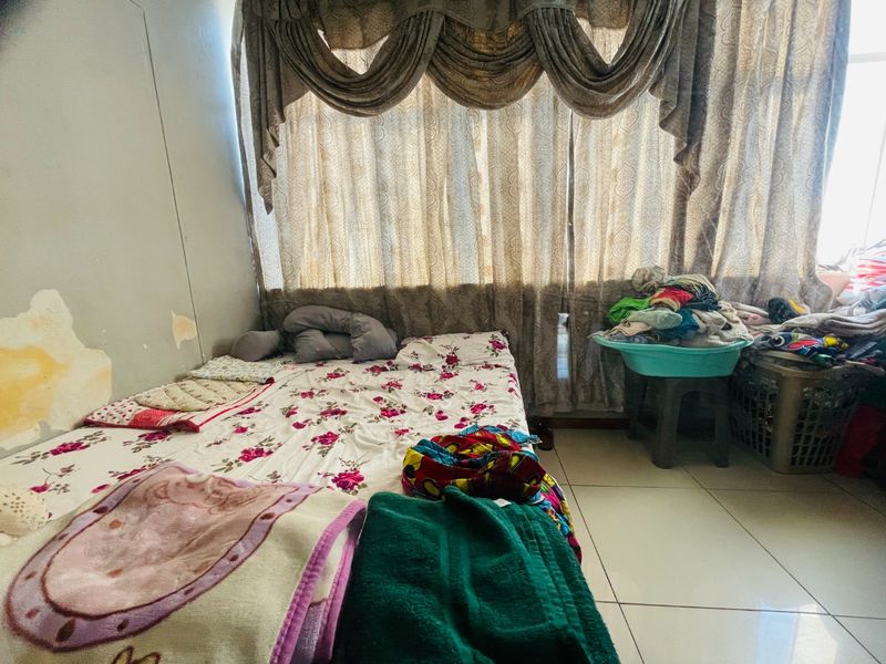 A bedroom featuring tiled flooring and large window curtains, currently showing signs of wall surface wear and peeling paint.