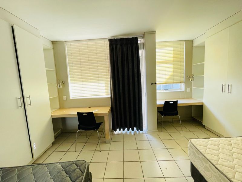 A functional bedroom featuring dual built-in desks, ample cupboard storage, tiled flooring, and large windows with venetian blinds for natural light.