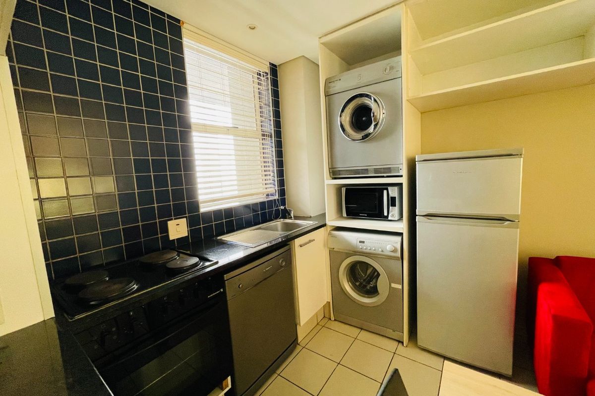 Compact kitchen featuring dark tiled backsplash, built-in oven and stove, integrated dishwasher, and dedicated laundry appliance housing with overhead shelving.