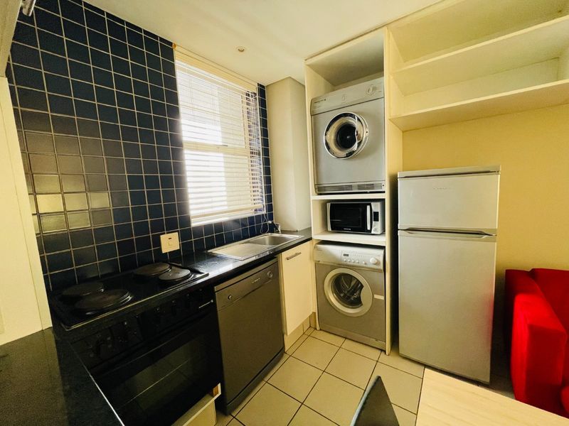 Compact kitchen featuring dark tiled backsplash, built-in oven and stove, integrated dishwasher, and dedicated laundry appliance housing with overhead shelving.