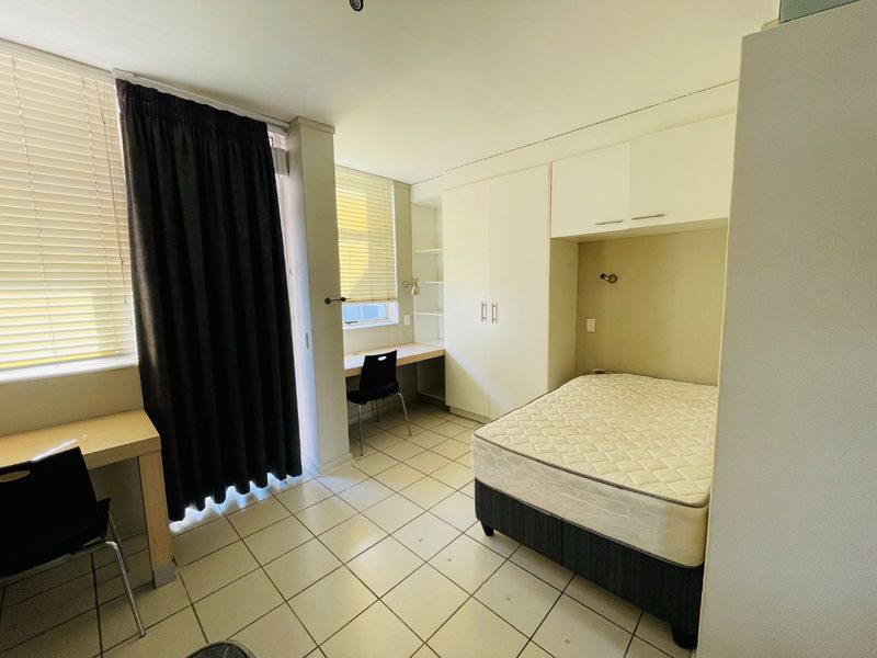 A functional bedroom featuring built-in cupboards, a dedicated study desk area, and tiled flooring with large windows for natural light.