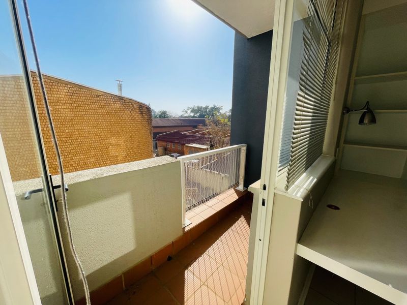 A sunny balcony featuring tiled flooring, a metal balustrade, and a built-in desk area with shelving, offering an open view of the surrounding neighborhood.
