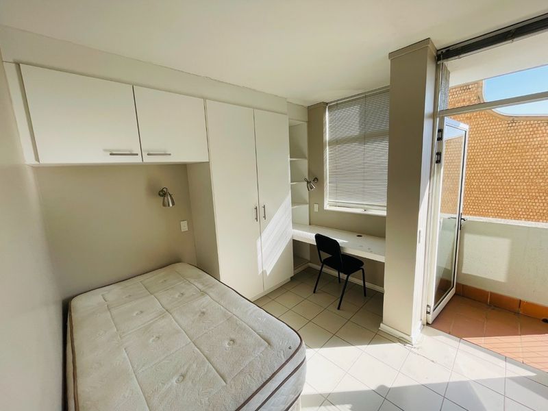 A bright bedroom featuring built-in cupboards, a dedicated study desk, tiled flooring, and direct access to a private balcony.