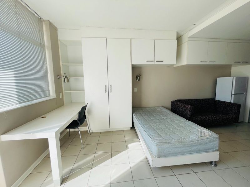 A functional bedroom featuring built-in cupboards, overhead storage, and a built-in desk with tiled flooring and natural light from the window.