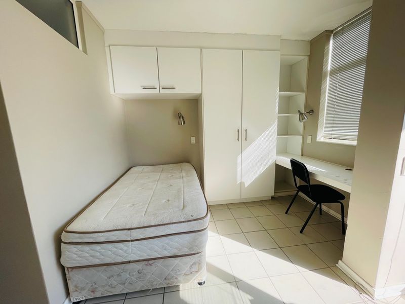 A functional bedroom featuring built-in wardrobes, overhead storage, and a dedicated built-in desk area with tiled flooring and natural light.