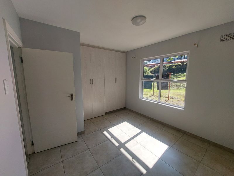 A bright, empty bedroom featuring neutral tiled flooring, built-in cupboards, and a large window providing natural light and a garden view.