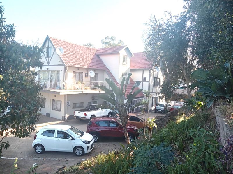 A multi-story apartment building featuring a tiled roof, balcony, and paved parking area, surrounded by established trees and landscaping.