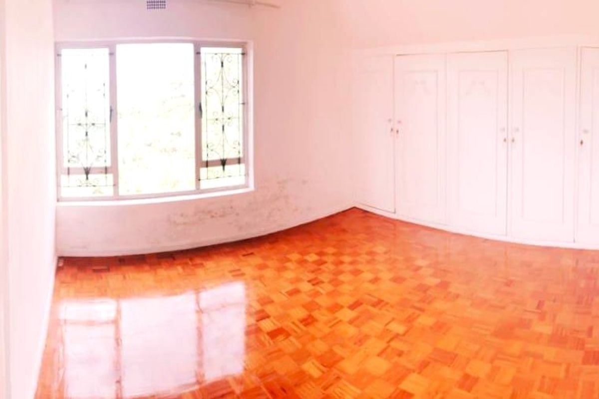 An empty bedroom featuring polished parquet flooring, built-in cupboards, and a window with security bars. The room shows signs of wall staining.