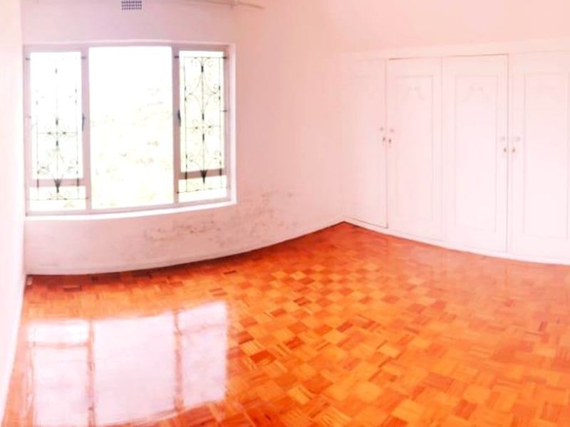 An empty bedroom featuring polished parquet flooring, built-in cupboards, and a window with security bars. The room shows signs of wall staining.