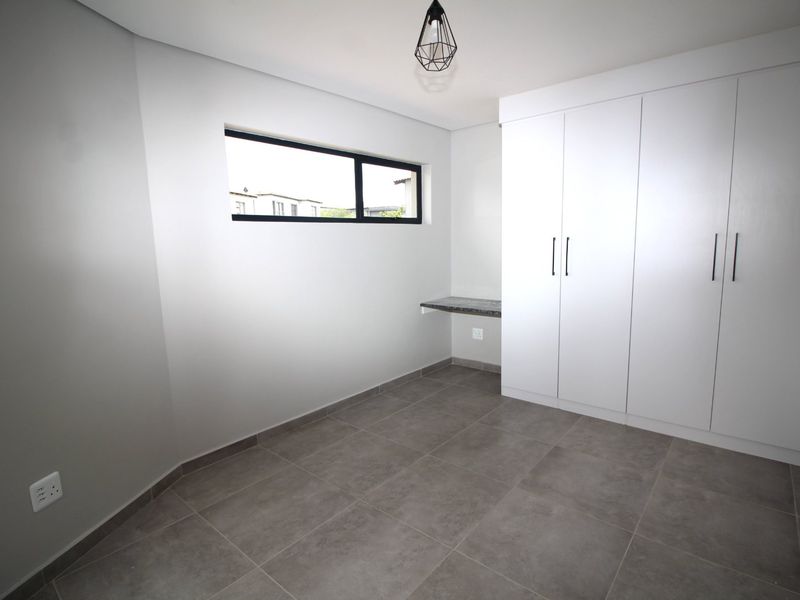 A clean, modern bedroom featuring neutral grey tiled floors, built-in white cupboards, a compact built-in desk, and a contemporary pendant light fixture.