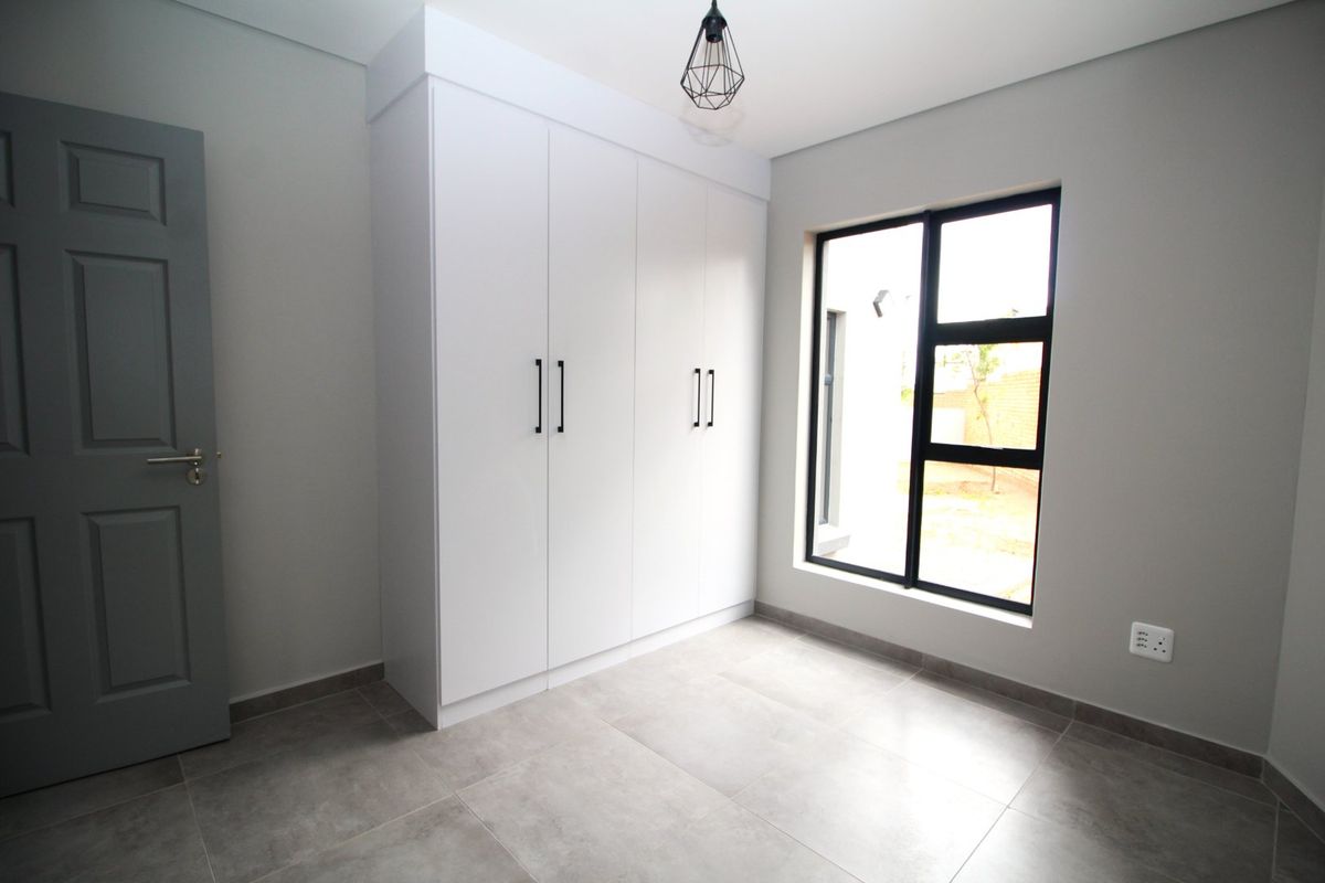A clean, modern bedroom featuring neutral grey walls, large format floor tiles, a built-in white wardrobe, and a contemporary black-framed window.
