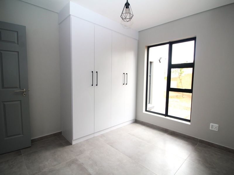 A clean, modern bedroom featuring neutral grey walls, large format floor tiles, a built-in white wardrobe, and a contemporary black-framed window.