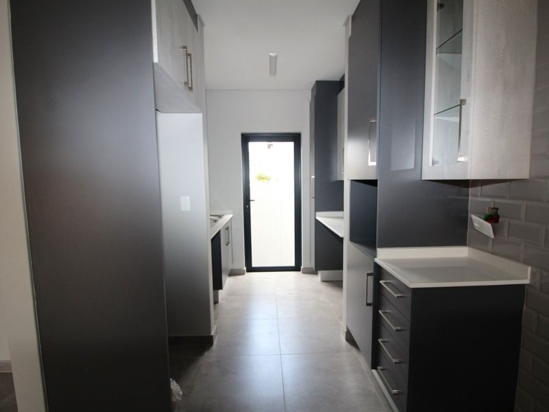 A modern, empty scullery featuring sleek dark cabinetry, stone countertops, subway tile backsplash, and tiled flooring with direct access to the exterior.