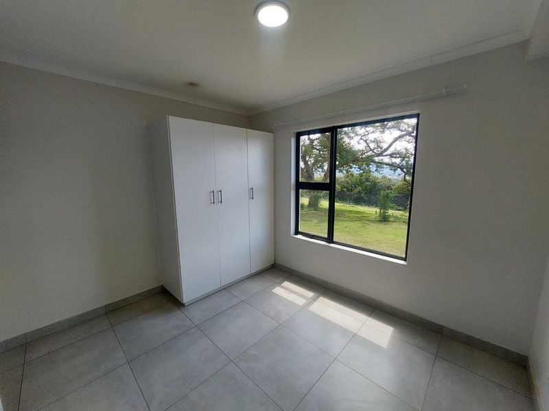 A bright, empty bedroom featuring neutral tiled flooring, a built-in wardrobe, and a large window with scenic garden views.