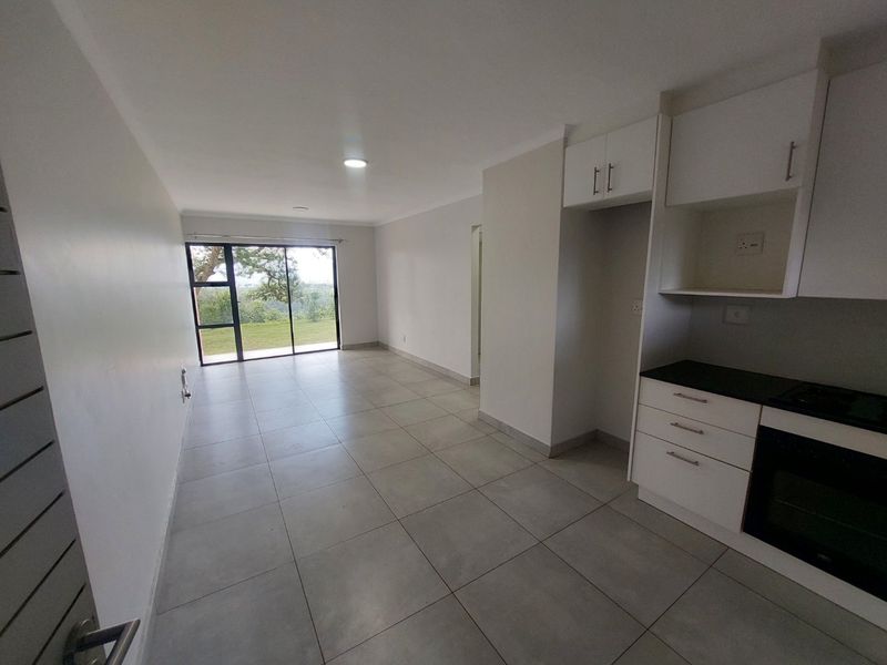 An open-plan living and kitchen area featuring modern white cabinetry, built-in oven, large format floor tiles, and a sliding door leading to an outdoor view.