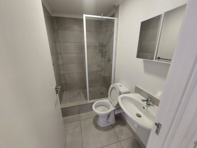 A clean, modern bathroom featuring a walk-in shower with glass enclosure, pedestal washbasin, and a wall-mounted mirror cabinet with neutral floor-to-ceiling tiling.
