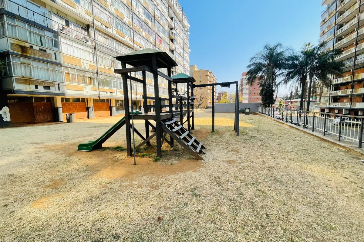 Communal outdoor area featuring a playground structure situated between apartment blocks with metal perimeter fencing and dry lawn.