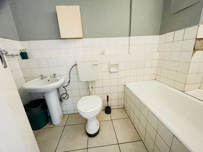 A functional bathroom featuring a pedestal basin, toilet, and bathtub with white wall tiling. The space requires cosmetic updates and repairs to finishes.
