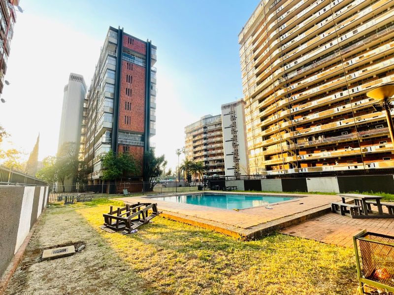 Communal outdoor pool area featuring a swimming pool, brick paved surround, and perimeter fencing within a high-rise apartment complex.