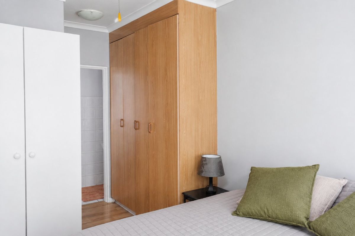 A bedroom featuring light wood-toned built-in wardrobes and neutral wall finishes, with an adjacent view into a tiled bathroom area.