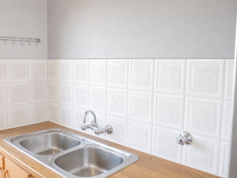 A clean kitchen area featuring a double stainless steel sink, wall-mounted faucet, laminate countertop, and white tiled backsplash.