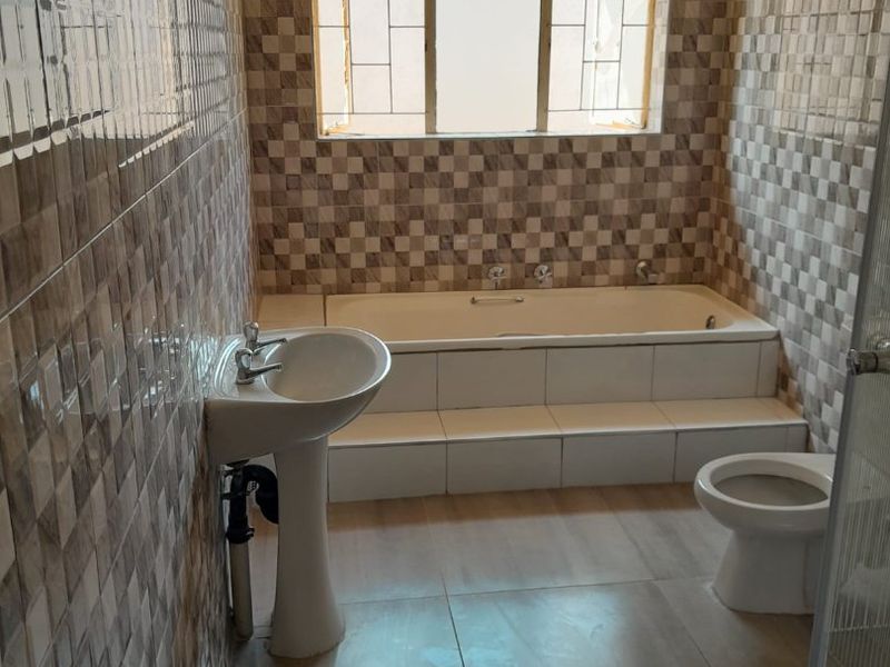 A functional bathroom featuring a built-in bathtub, pedestal basin, and toilet, finished with neutral checkered wall tiling and floor tiles.
